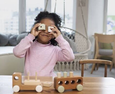 Imagem de uma criança brincando com peças, nas duas mãos, cada uma tem uma pecinha e ela coloca na altura dos olhos, em cima da mesa, brinquedos e atrás dela um balanço.