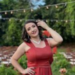 Imagem de uma mulher, usando vestido de festa vermelho, maquiada e com uma das mãos proximo do cabelo. Ao fundo lampadas em um varal e bastante árvores verdes.