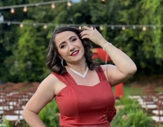 Imagem de uma mulher, usando vestido de festa vermelho, maquiada e com uma das mãos proximo do cabelo. Ao fundo lampadas em um varal e bastante árvores verdes.