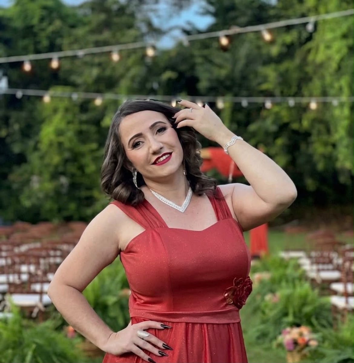Imagem de uma mulher, usando vestido de festa vermelho, maquiada e com uma das mãos proximo do cabelo. Ao fundo lampadas em um varal e bastante árvores verdes.