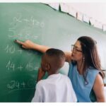 Imagem de uma professora, ensinando matemática ao aluno no quadro verde. A professora usa óculos e aponta para a soma no quadro.