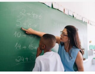 Imagem de uma professora, ensinando matemática ao aluno no quadro verde. A professora usa óculos e aponta para a soma no quadro.