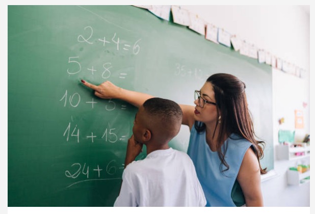 Imagem de uma professora, ensinando matemática ao aluno no quadro verde. A professora usa óculos e aponta para a soma no quadro.