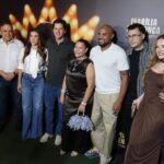 Imagem do governador, sua esposa e amigos pousando para fotos. Atrás um painel lançamento de série documental sobre Marília Mendonça
