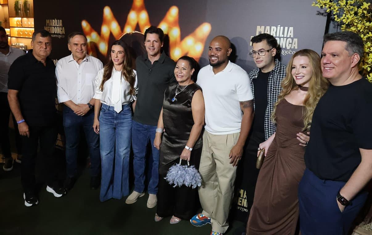 Imagem do governador, sua esposa e amigos pousando para fotos. Atrás um painel lançamento de série documental sobre Marília Mendonça