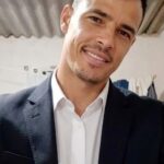 Imagem de um homem, usa camisa branca e blazer preto, sorri para a foto.