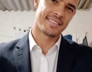 Imagem de um homem, usa camisa branca e blazer preto, sorri para a foto.
