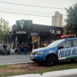 Imagem da cena do crime, onde aparece uma distribuidora ao fundo, pessoas em pé, próximoa porta principal da distribuidora, carro da polícia militar e fita zebrada.