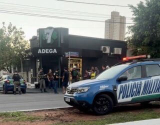 Imagem da cena do crime, onde aparece uma distribuidora ao fundo, pessoas em pé, próximoa porta principal da distribuidora, carro da polícia militar e fita zebrada.