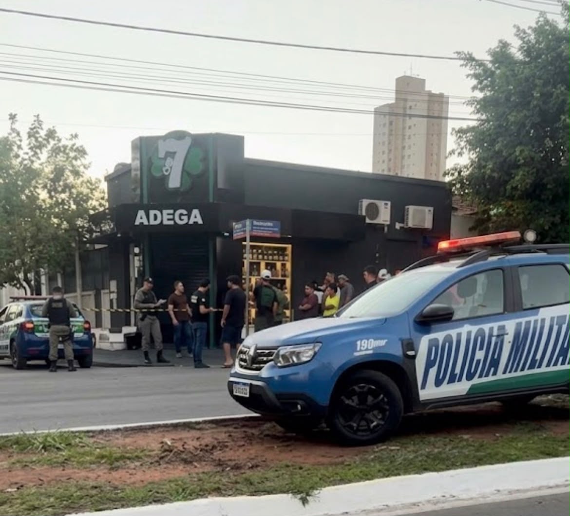 Imagem da cena do crime, onde aparece uma distribuidora ao fundo, pessoas em pé, próximoa porta principal da distribuidora, carro da polícia militar e fita zebrada.