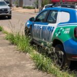 Imagem de uma viatura da Polícia Militar estacionando na porta de uma casa. Na calçada alguns matinhos e a frente outro carro estacionado.
