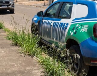 Imagem de uma viatura da Polícia Militar estacionando na porta de uma casa. Na calçada alguns matinhos e a frente outro carro estacionado.