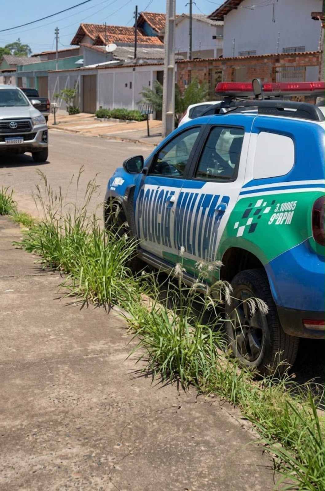 Imagem de uma viatura da Polícia Militar estacionando na porta de uma casa. Na calçada alguns matinhos e a frente outro carro estacionado.