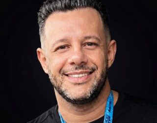 Imagem de uma homem com camiseta preta, cordão azul no pescoço, cabelos grisalhos e barba, olhando e sorrindo para a foto.