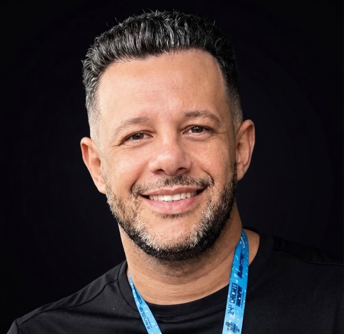 Imagem de uma homem com camiseta preta, cordão azul no pescoço, cabelos grisalhos e barba, olhando e sorrindo para a foto.