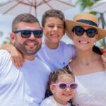 Imagem de Amilton Filho com sua esposa Carol e dois filhos, usando roupas brancas e óculos de sol.