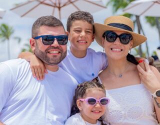 Imagem de Amilton Filho com sua esposa Carol e dois filhos, usando roupas brancas e óculos de sol.