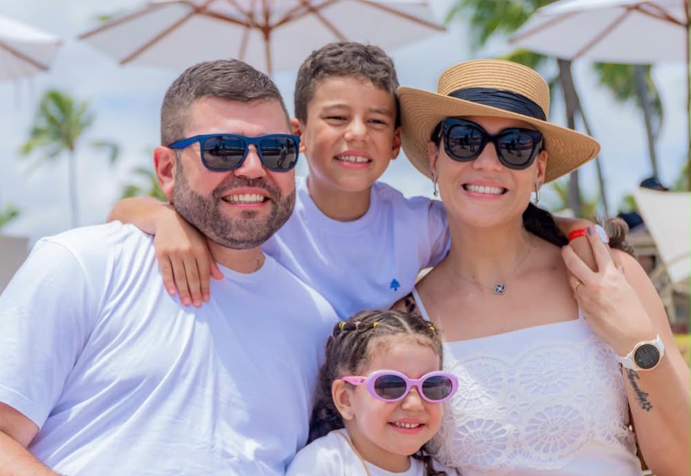 Imagem de Amilton Filho com sua esposa Carol e dois filhos, usando roupas brancas e óculos de sol.