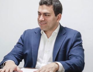 Imagem de José Frederico Lyra Netto, sentado, com as mãos postas em cima da mesa, algumas folhas em cima da mesa. Ele está usando terno sem gravata, olhando para o lado e sorrindo.