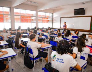 Imagem de uma sala de aula com vários alunos da escola pública, utilizando uniforme. As carteiras são brancas e azuis e há uma professora a frente da turma.