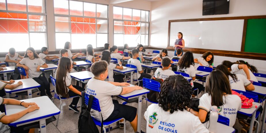 Imagem de uma sala de aula com vários alunos da escola pública, utilizando uniforme. As carteiras são brancas e azuis e há uma professora a frente da turma.