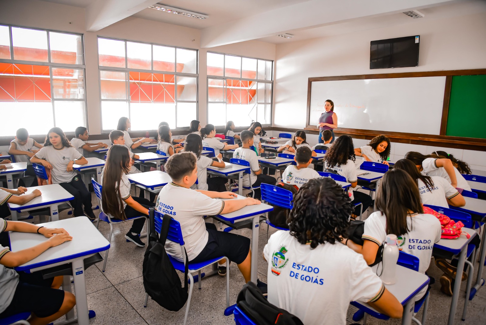Imagem de uma sala de aula com vários alunos da escola pública, utilizando uniforme. As carteiras são brancas e azuis e há uma professora a frente da turma.